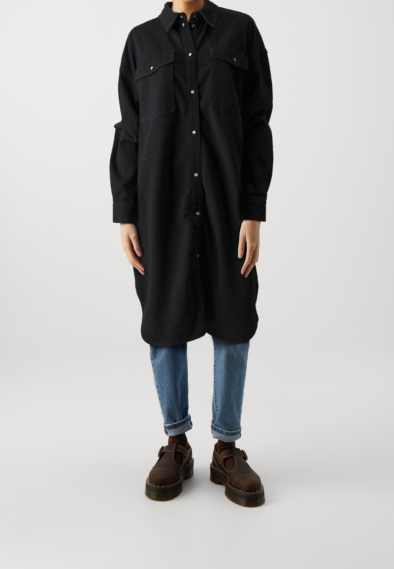 Personne portant une longue robe-chemise noire boutonnée, un jean bleu clair retroussé et des chaussures épaisses marron foncé sur un fond clair uni.