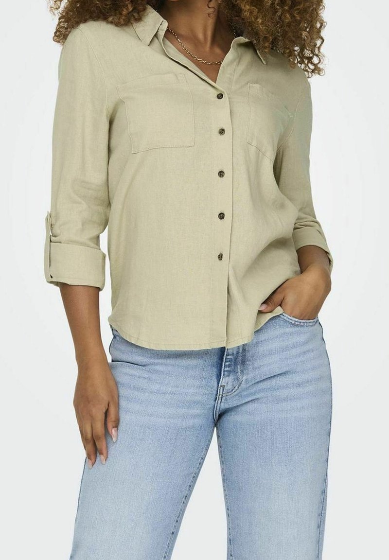 Lys olivengrøn knap-op skjorte med to brystlommer, opkrøllede ærmer og brune knapper, parret med blå jeans. Glat stoftekstur.