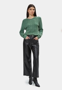 Maglione verde a grana di riso con orlo a coste abbinato a pantaloni a gamba larga neri in ecopelle e stivaletti neri con punta. Dettaglio della cintura argentata incluso.
