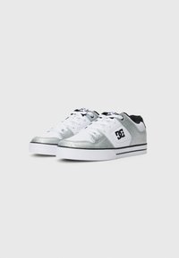 Zapatillas bajas blancas y plateadas con logo negro en el lateral, suelas gruesas blancas y cordones blancos, exhibidas sobre un fondo claro y sencillo.