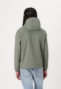 Jachetă ușoară de culoare verde olive, cu glugă, manșete elastice și croială lejeră, purtată peste blugi albaștri deschiși. Textură netedă, design simplu.