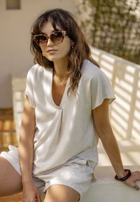 Femme portant des lunettes de soleil à monture écaille et une chemise à manches courtes beige clair avec un short, assise sur un rebord blanc extérieur avec une lumière du soleil tamisée.