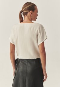 Weißes kurzärmeliges Blusenshirt mit glatter Textur, kombiniert mit einem schwarzen Lederröck mit seitlichen Druckknopfdetails, von hinten betrachtet.