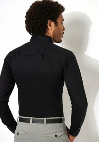 Zwarte langetermijnshirt met een strakke pasvorm en subtiele textuur, voorzien van een scherpe kraag en contrasterende knopen. G gedragen met lichtgrijze broek.