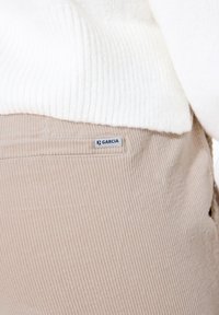 Close-up van beige corduroy broek met een klein "Garcia" label genaaid nabij de achterzak, gedragen met een witte ribbeltrui.