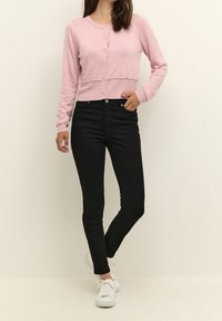 Vaaleanpunainen lyhyt neulejakku pyöreällä pääntiellä ja napituksella, yhdistettynä korkeavyötäröisiin mustiin farkkuihin ja valkoisiin tennareihin. Sileä pinta.