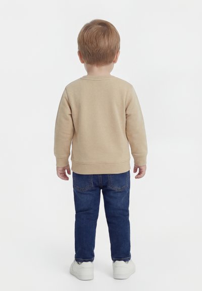 Jeune enfant aux cheveux blonds portant un sweat-shirt beige, un jean bleu et des baskets blanches, debout de dos sur fond blanc.