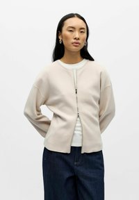 Veste zippée beige clair en tissu doux, avec épaules tombantes et encolure arrondie. Portée sur un haut blanc et associée à un jean foncé.