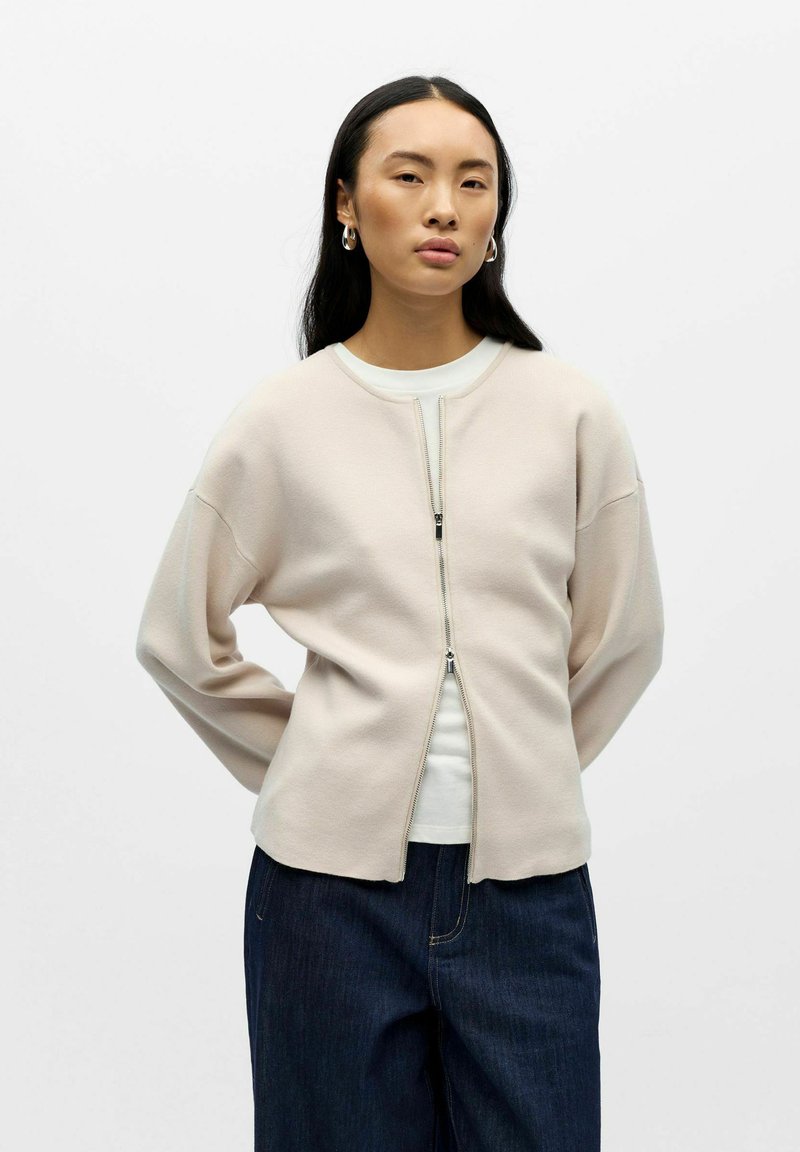 Veste zippée beige clair en tissu doux, avec épaules tombantes et encolure arrondie. Portée sur un haut blanc et associée à un jean foncé.