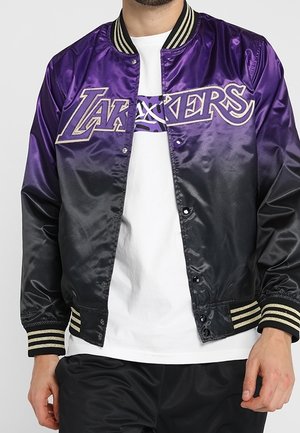 Persona vistiendo una chaqueta satinada morada y negra de Los Angeles Lakers sobre una camisa blanca y pantalones negros, de pie frente a un fondo liso.