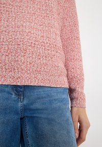 Gros plan sur une personne portant un pull en tricot rose moucheté et un jean en denim bleu, avec une main détendue le long du corps.
