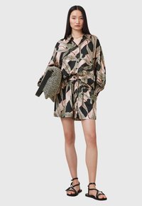 Zwarte oversized shirtjurk met kleurrijk bladerenpatroon, taillering en korte mouwen; gecombineerd met een gestreepte handtas en zwarte sandalen.