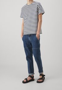 Gestreepte katoenen T-shirt in marineblauw en crème, gecombineerd met blauwe spijkerbroek met zichtbare naden en opgerolde mouwen. Zwarte platte sandalen.