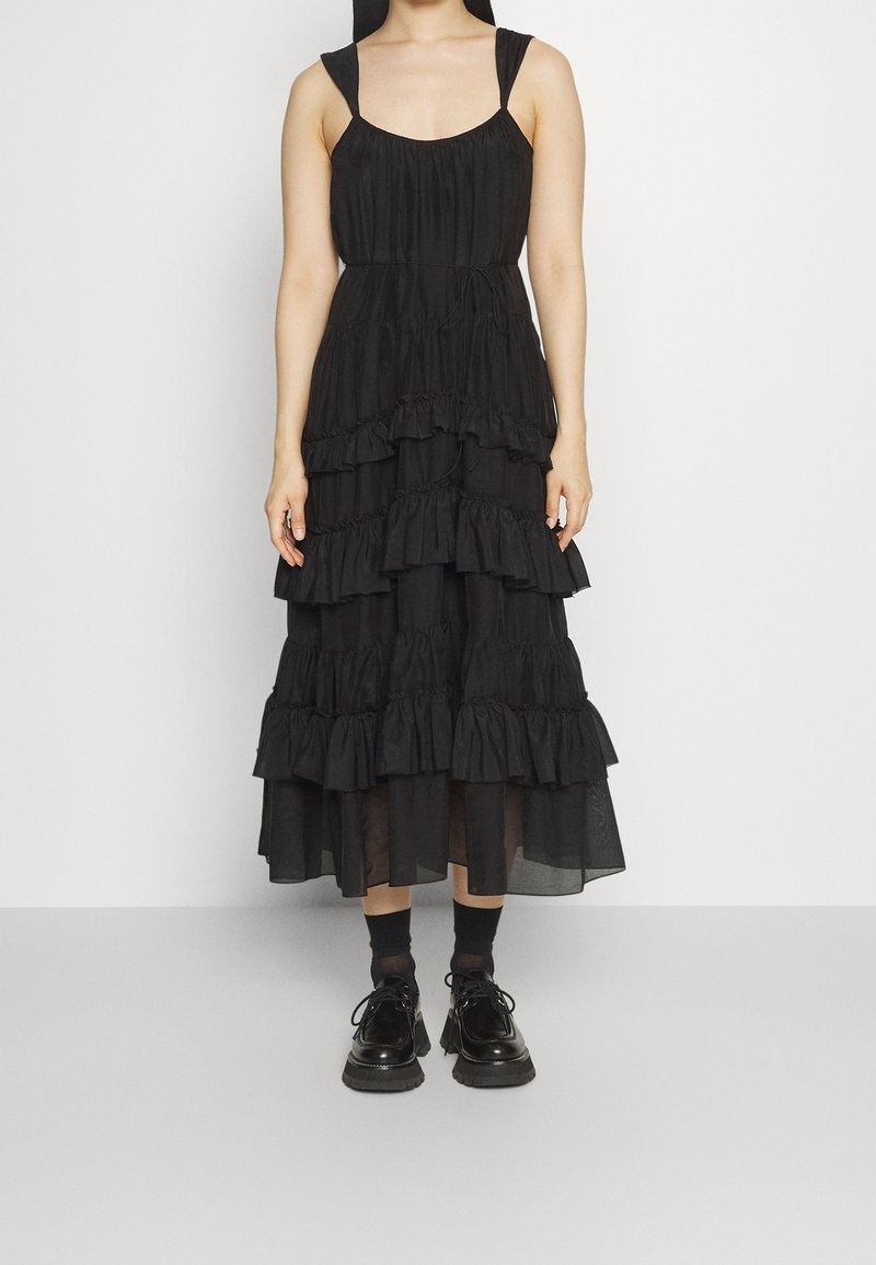 Femme portant une robe midi noire sans manches à volants superposés avec de larges bretelles épaisses et des chaussures noires à lacets avec semelles épaisses.