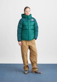 Padded green jacket with a high collar and front zipper. Paired with beige pants and gray shoes. Minimalistic design, no visible patterns.