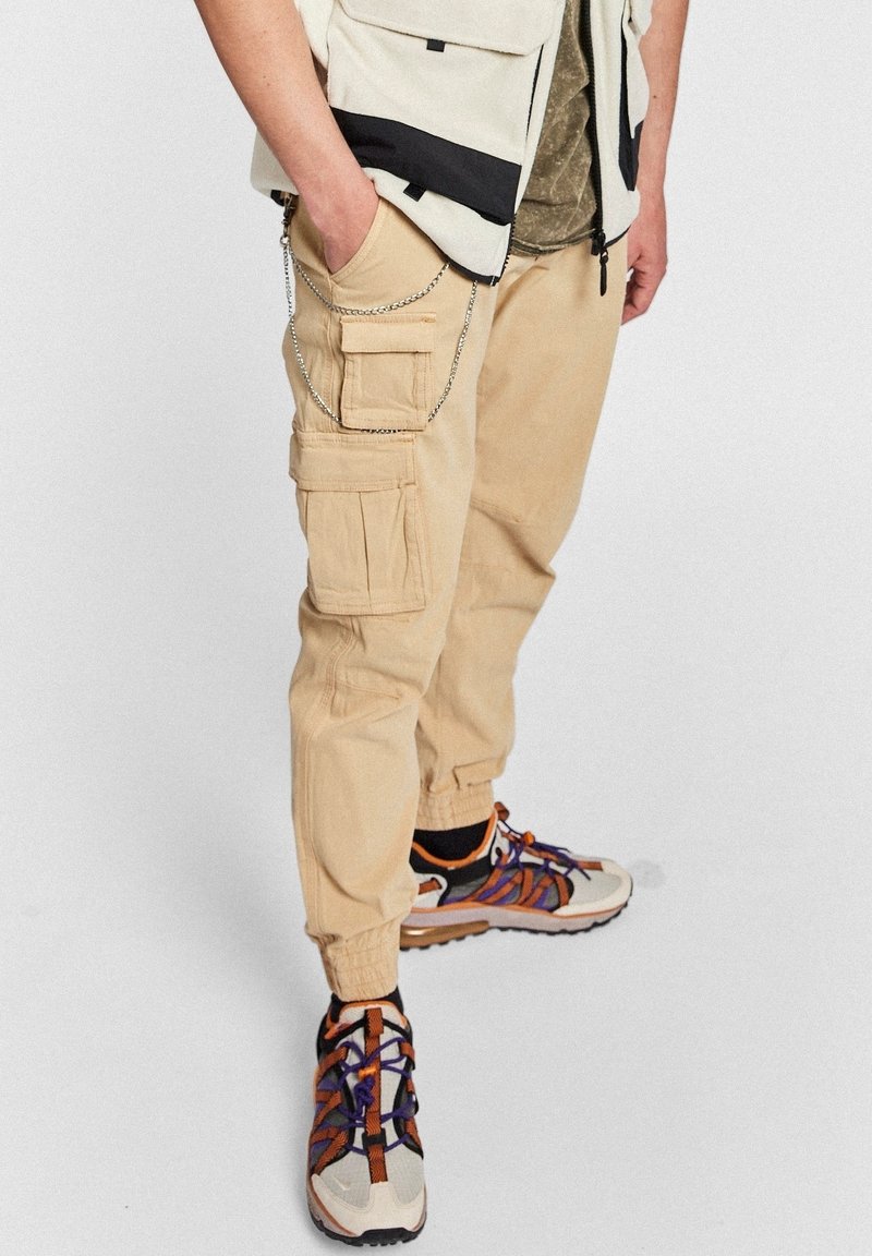 Persoon in beige cargobroek met kettingdetail, witte en oranje sneakers met paarse veters, en een gedeeltelijk zicht op een witte vest.