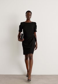 Abito nero a maniche corte con scollatura quadrata, forma aderente e dettagli arricciati sulle maniche. Indossato con una pochette nera e tacchi alti.