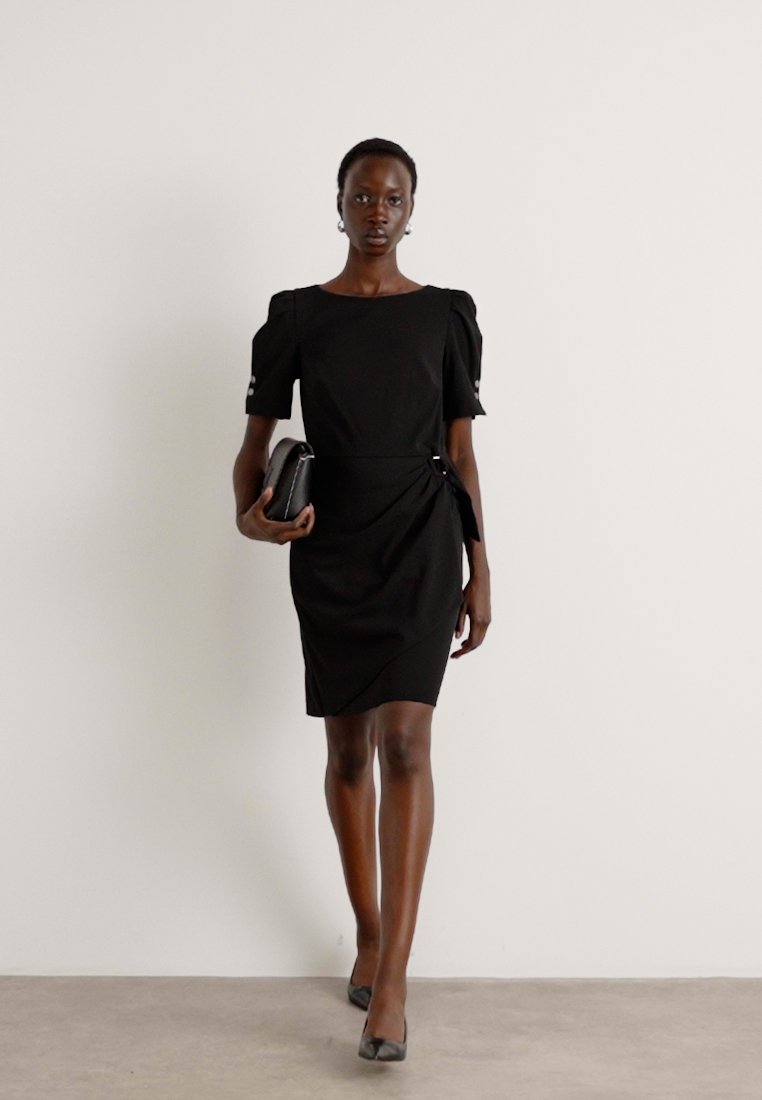 Robe noire à manches courtes avec décolleté carré, silhouette ajustée et détails froncés aux manches. Portée avec une pochette noire et des talons.