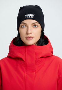 Berretto lavorato a maglia nero con logo "rukka"; giacca a hood rossa brillante con texture liscia e colletto alto; sfondo chiaro.