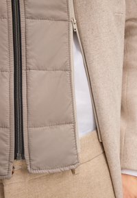 Jasnobeżowa pikowana kamizelka z czarnym zamkiem błyskawicznym, noszona pod jasnoszarym dopasowanym płaszczem, zestawiona z beżowymi materiałowymi spodniami.