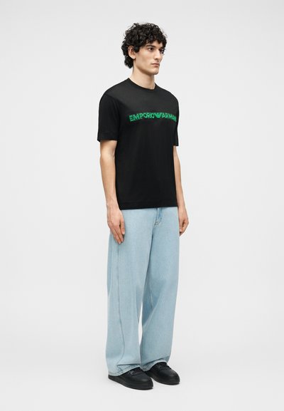 Άνδρας με σγουρά μαλλιά που φοράει μαύρο t-shirt Emporio Armani, ανοιχτό μπλε τζιν φαρδιάς γραμμής και μαύρα αθλητικά παπούτσια, στέκεται μπροστά σε απλό φόντο.