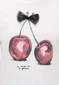 T-shirt blanc avec une illustration noire et rouge de deux cerises, avec un accent en nœud en mesh noir au-dessus. Le texte indique : "la cerise sur le gâteau."