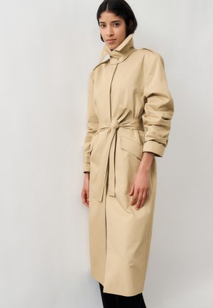 Donna con capelli scuri che indossa un trench beige con cintura, collo alto e maniche lunghe, in piedi davanti a uno sfondo chiaro e semplice.