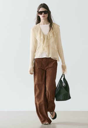 Femme portant de grandes lunettes de soleil, un chemisier en dentelle crème, un pantalon large marron, des tongs noires, et tenant un sac à main noir, marchant vers l'avant.