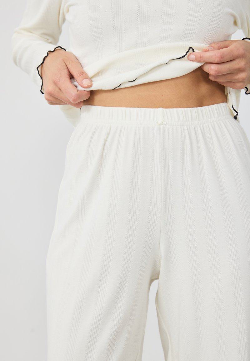 Ensemble loungewear en tissu côtelé blanc avec une taille élastique et des bords noirs festonnés le long des poignets. Texture douce avec des lignes verticales.