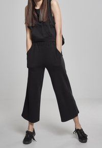 Zwarte mouwloze top en wijde zwarte broek van zacht materiaal, met een elastische tailleband en zijzakken, gecombineerd met zwarte sneakers.