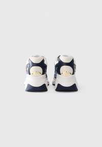 Un paio di sneakers bianche e blu navy viste da dietro su uno sfondo bianco semplice, che mostrano suole spesse e dettagli del logo.