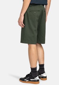 Gr�üne Baumwollshorts mit geradem Schnitt, Gesäßtaschen und Knopfverschluss. Getragen mit schwarzen Turnschuhen und dunklen Socken.