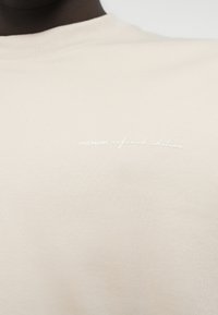 Μπεζ φούτερ με στρογγυλή λαιμόκοψη από απαλό, υφασμάτινο υλικό με υφή· διαθέτει το λεκτικό «PREMIUM refined edition» σε λευκό, ελαφρύ σχεδιασμό.