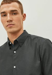 Camisa negra de botones con un acabado texturizado, que presenta un cuello clásico y botones blancos. El interior del cuello tiene un acento estampado en azul.