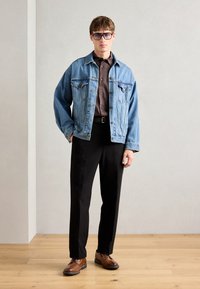 Denim jacket in light blue, brown button-up shirt, black trousers, brown shoes. Model stands against a neutral background, hands relaxed.