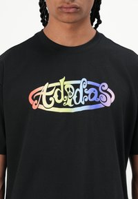 Črna bombažna majica s pisanim logotipom "Adidas" s stiliziranimi črkami. Kratki rokavi in izrez z okroglim ovratnikom.