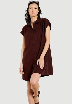 Žena v tmavohnedej krátkorukávovej šatách s gombíkmi vpredu, slnečné okuliare na hlave, stojí s jednou nohou cez druhú.