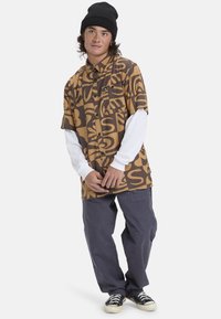 Korte mouw blouse in bruin met zwarte abstracte patronen, gecombineerd met een witte lange mouw shirt en grijze broek. Zwarte beanie.