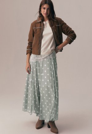 Donna che indossa una giacca marrone borchiata, un top bianco con rifiniture in pizzo, una gonna verde chiaro a pois e stivaletti marroni, in piedi su uno sfondo neutro.