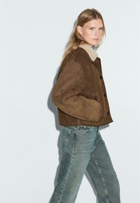 Bruine suede jas met een schapenvachtkraag, met een rechte pasvorm, knopen aan de voorkant en zijzakken, gecombineerd met vervaagde blauwe jeans.