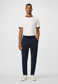 Witte t-shirt met bordeauxrode rand, gecombineerd met donkerblauwe sweatpants met zijzakken en trekkoord. Witte sneakers maken de outfit compleet.