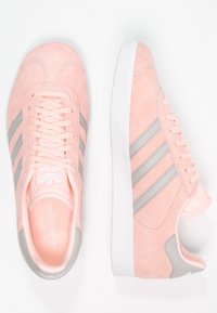 Růžové semišové tenisky s třemi horizontálními šedými pruhy, texturovanou gumovou podrážkou a polstrovaným límcem. Branding je viditelný na jazýčku a stélce.