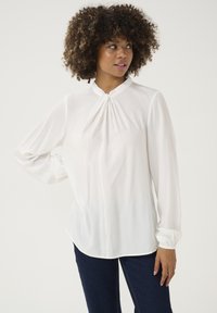 Blusa bianca a maniche lunghe con scollatura arricciata, vestibilità leggermente loose e texture morbida e liscia. Abbinata a jeans scuri.