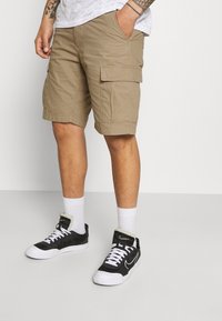 Shorts cargo beiges avec deux poches latérales, une texture lisse et des ourlets cousus. Portés avec des baskets noires et des chaussettes blanches au niveau des chevilles.