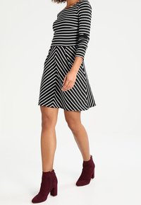 Robe à rayures noires et blanches avec des manches longues et une jupe évasée, associée à des bottines bordeaux. La robe présente des motifs en diagonale.