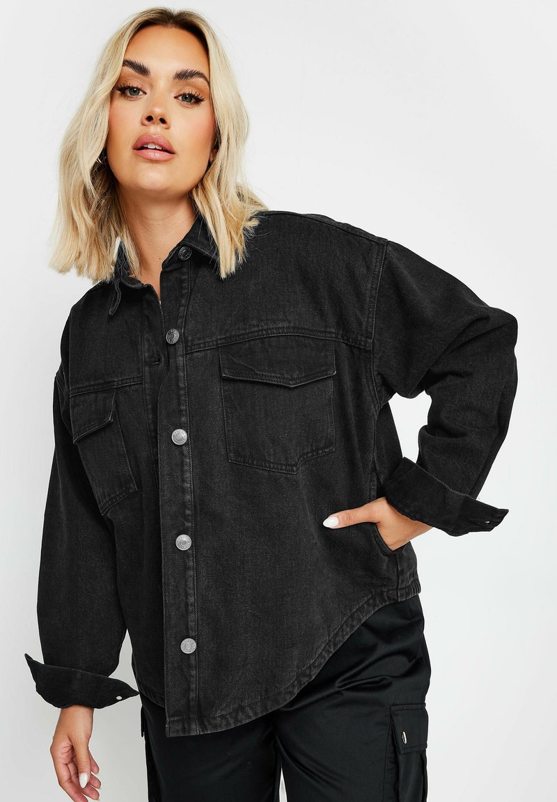 Musta farkkupaita-takki, jossa on napitettu etuosa, rintataskut ja hieman väljä muotoilu. Kangas on sileäpintainen ja tekstuuri on näkyvissä.