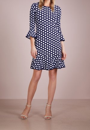 Femme portant une robe bleu marine longueur genou à pois blancs avec des manches trois-quarts volantées, assortie à des talons clairs à bride chevillée.
