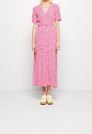 Femme portant une robe portefeuille rose et blanche à motifs, manches courtes, et des sabots beige, debout devant un fond blanc uni.