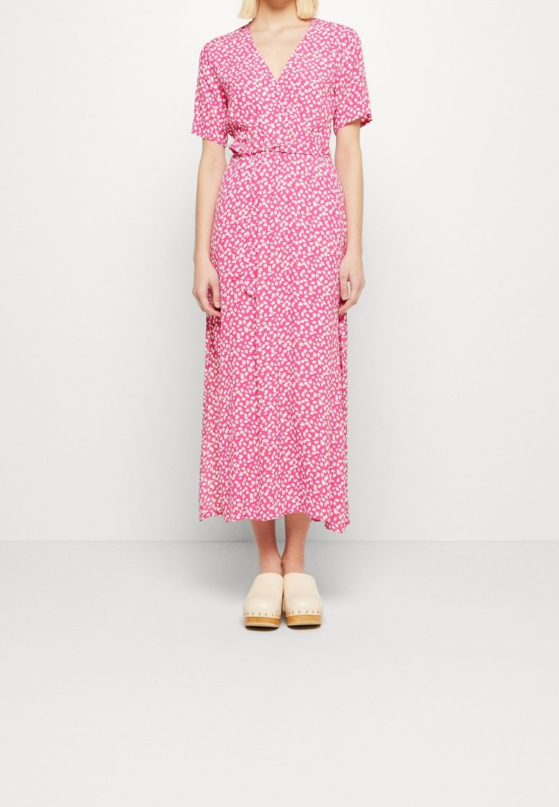 Femme portant une robe portefeuille rose et blanche à motifs, manches courtes, et des sabots beige, debout devant un fond blanc uni.