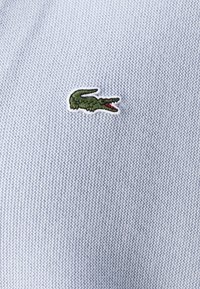 Světle modrá pletená polokošile s vyšívaným zeleným krokodýlím logem s červenými akcenty. Detailní pohled na strukturu látky.